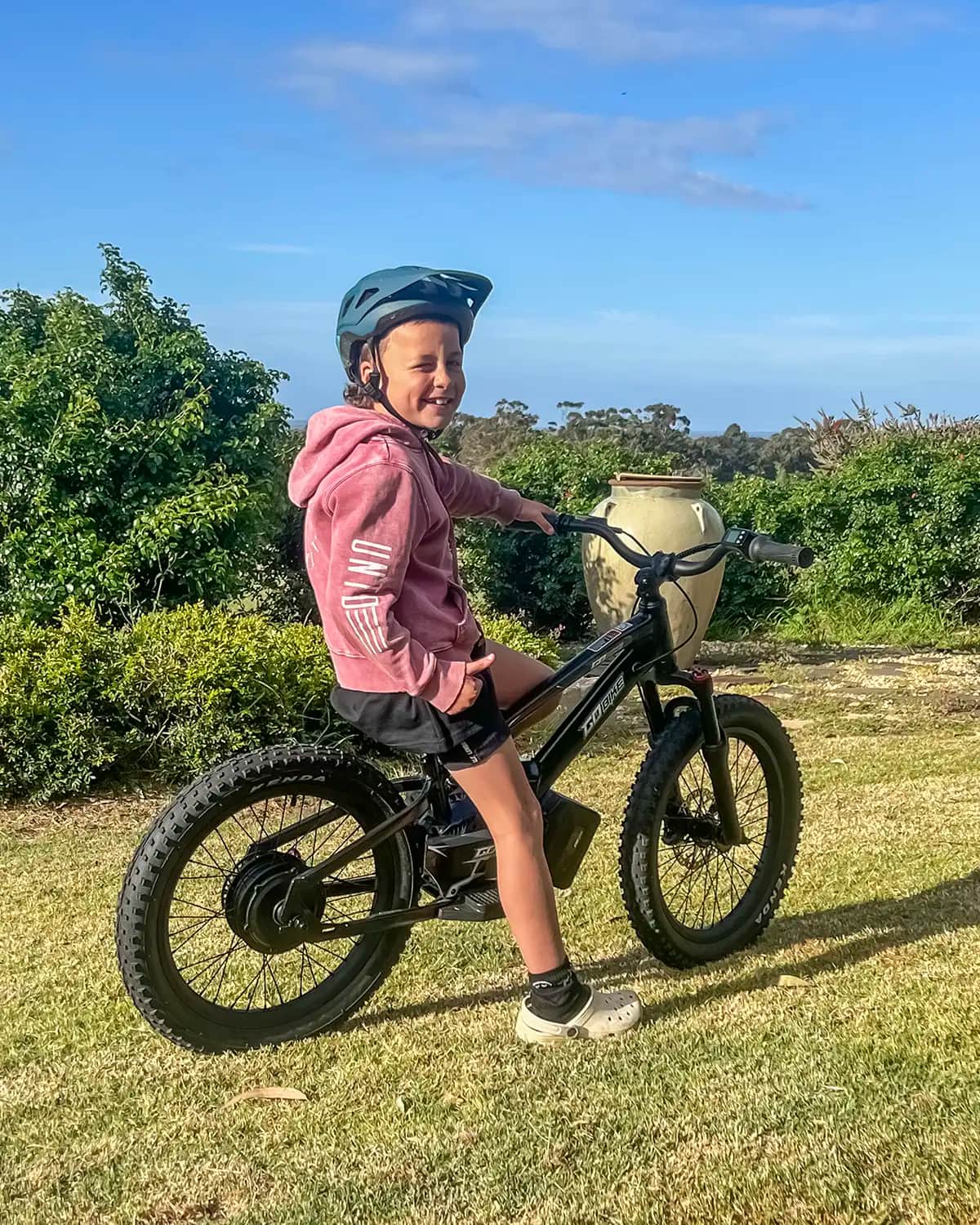 Smiling girl in a helmet and pink hoodie sitting on her GoBike 20 in the backyard