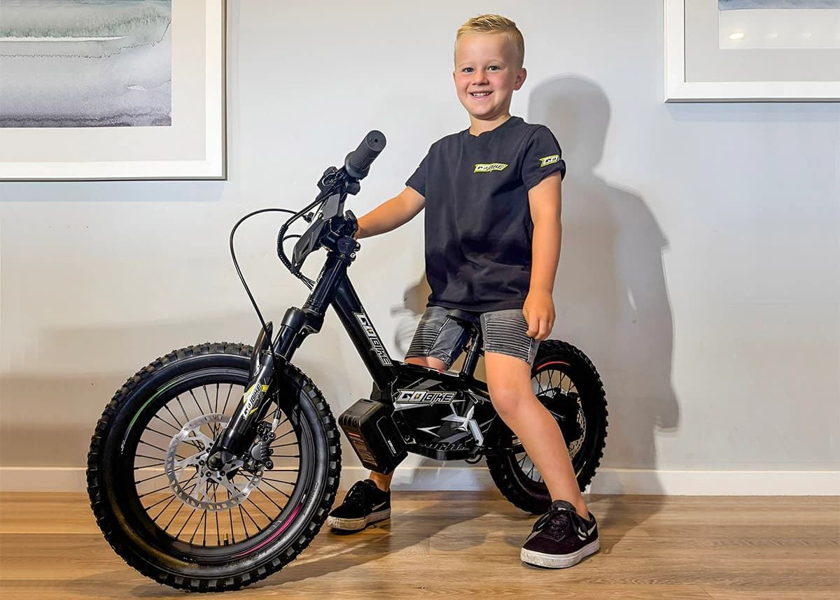 A proud young boy smiling with his new Gobike 16 electric dirt bike indoors.