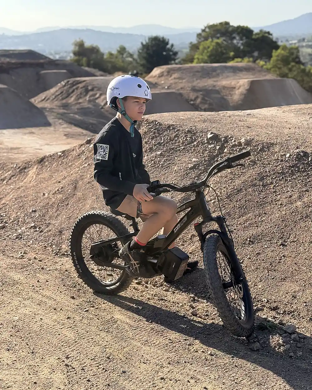 Boy resting on his GoBike kids electric motorbike at a dirt jump park in late afternoon light
