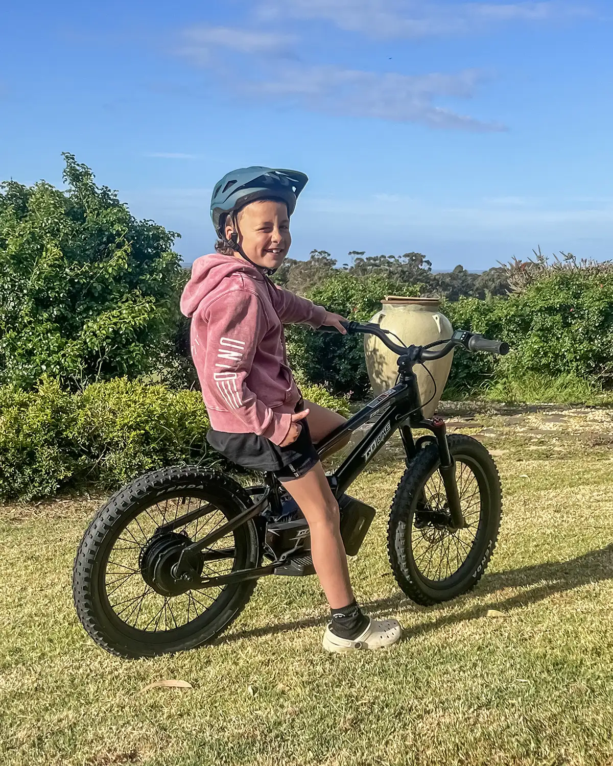 Smiling girl in a helmet and pink hoodie sitting on her GoBike 20 electric bike for kids in the backyard