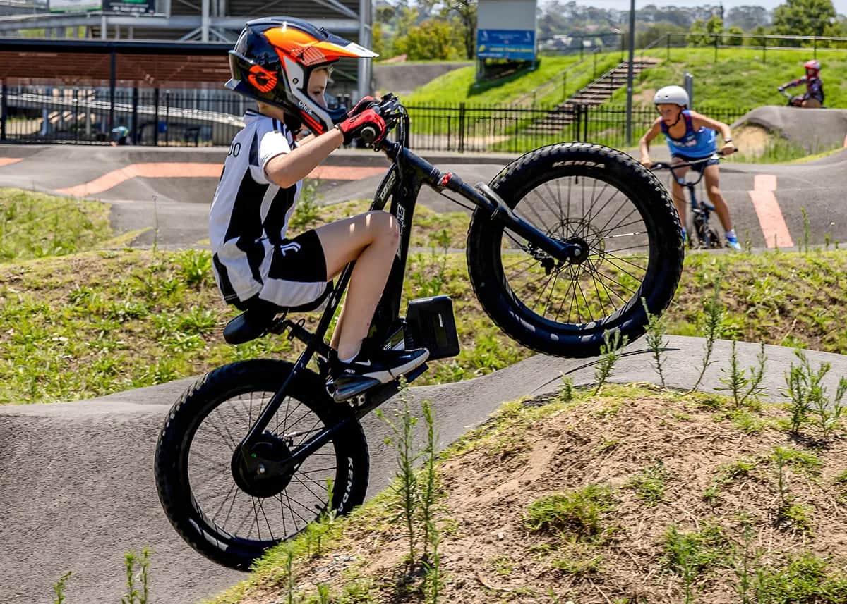 Kid in an orange helmet popping a wheelie on his GoBike 20 electric dirt bike at a pump track