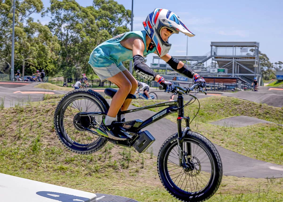 Kid in a stars-and-stripes helmet catching air on a GoBike 20 kids electric dirt bike at a bike park