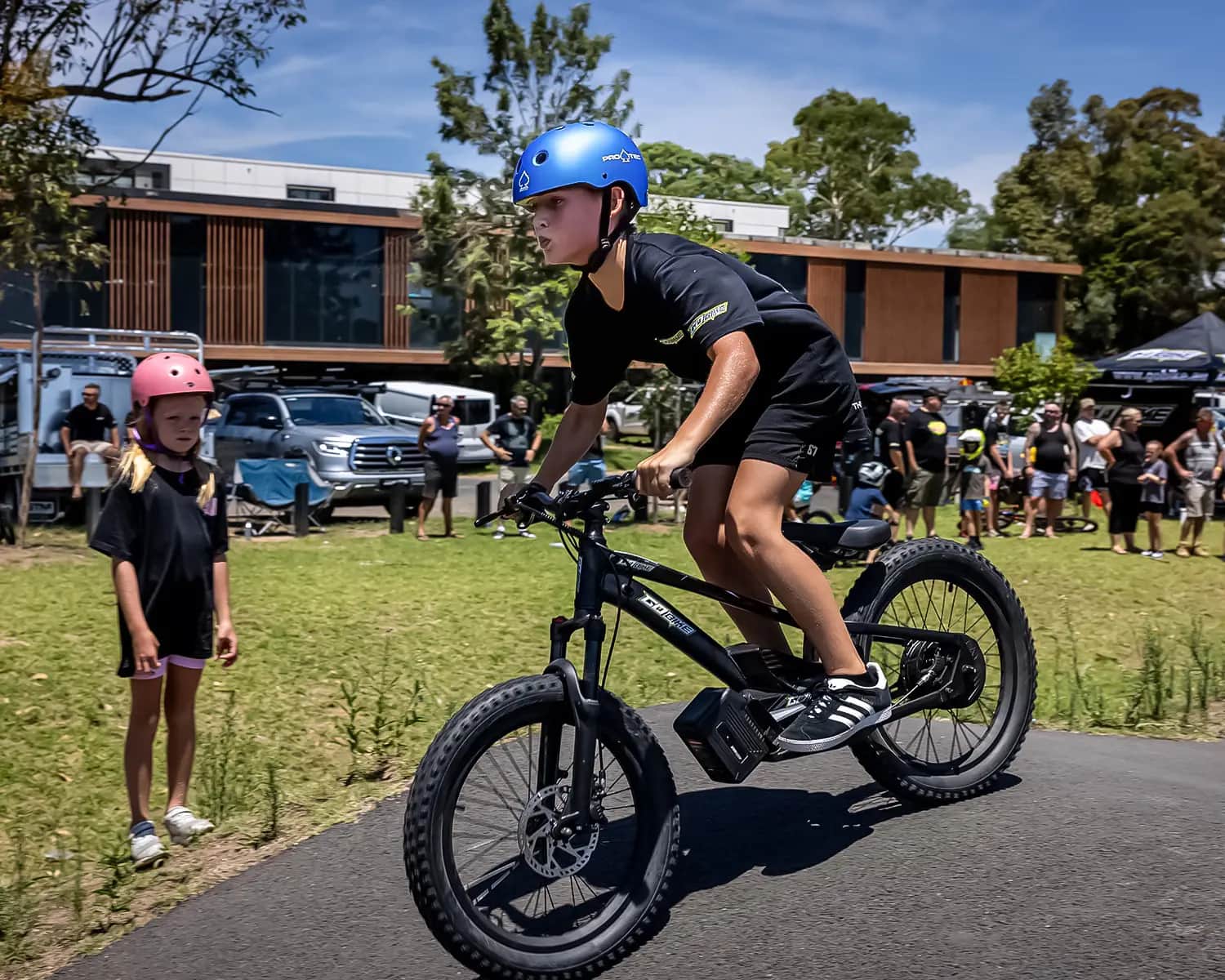 Teenager in a blue helmet riding a GoBike 20 inch electric bike on a pump track as families watch