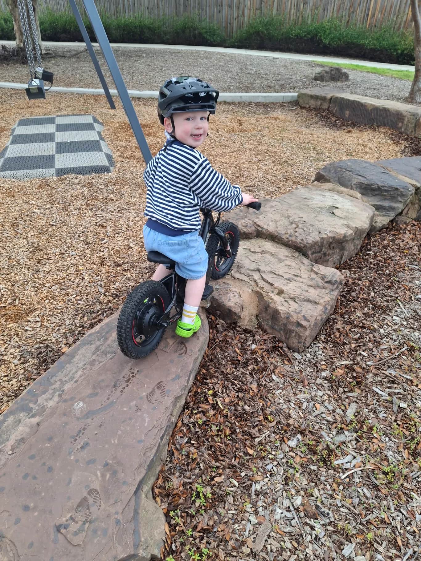 Smiling toddler in a black helmet and striped top riding his GoBike 12 over rocks at a playground