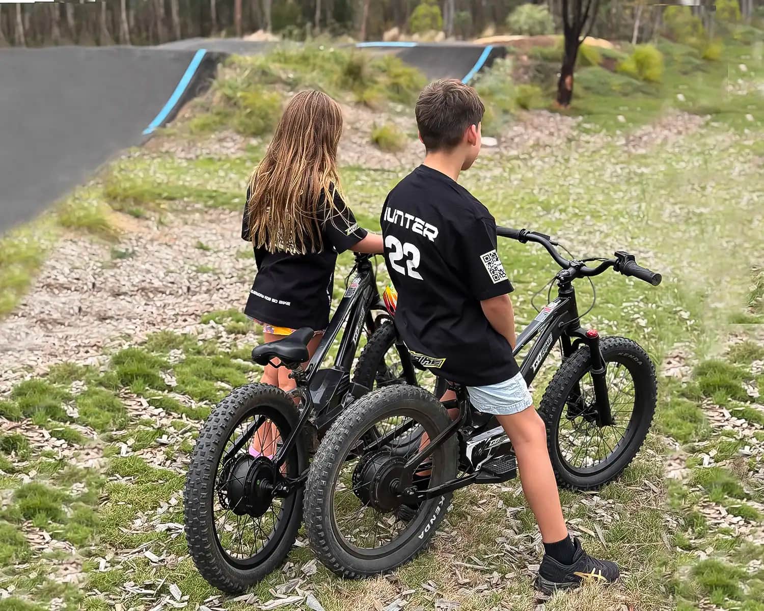 Two kids in GoBike riding jerseys on their GoBike 20 inch electric bikes, looking out at a pump track